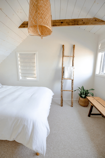 Chambre lumineuse avec lit blanc, plafond incliné en bois, échelle, plante, banc et abat-jour osier.