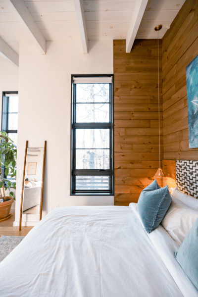 Chambre moderne avec mur en bois, lit blanc, coussins bleu clair, fenêtre, miroir, plante verte.